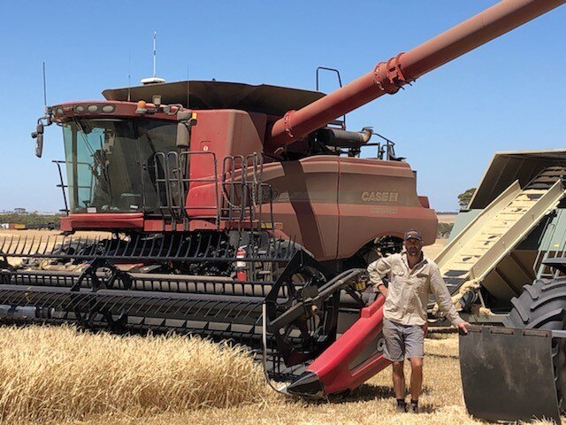 A man stands in front of a header at rest in a golden field.