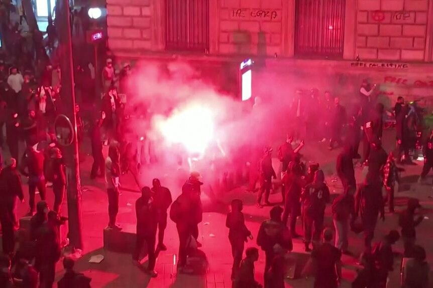 A crowd of people on the street with a red-tinted gas emitting from a flare.