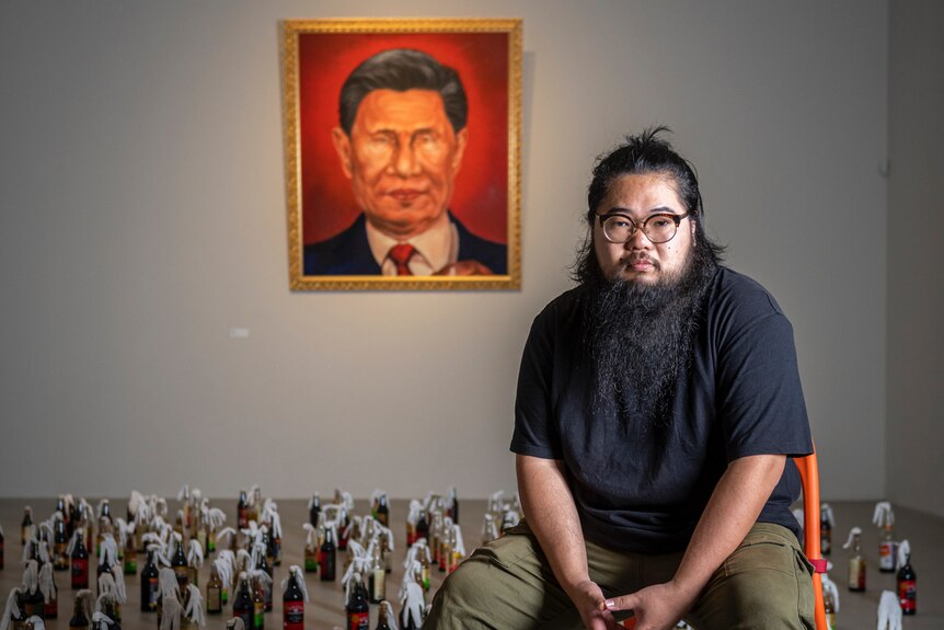 Badiucao in front of artwork, which depicts Chinese official and smaller art work pieces on the floor, wearing masks.