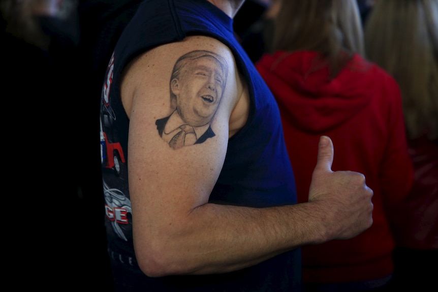On a man's upper arm, a black and white tattoo of Trump's face can be seen. 