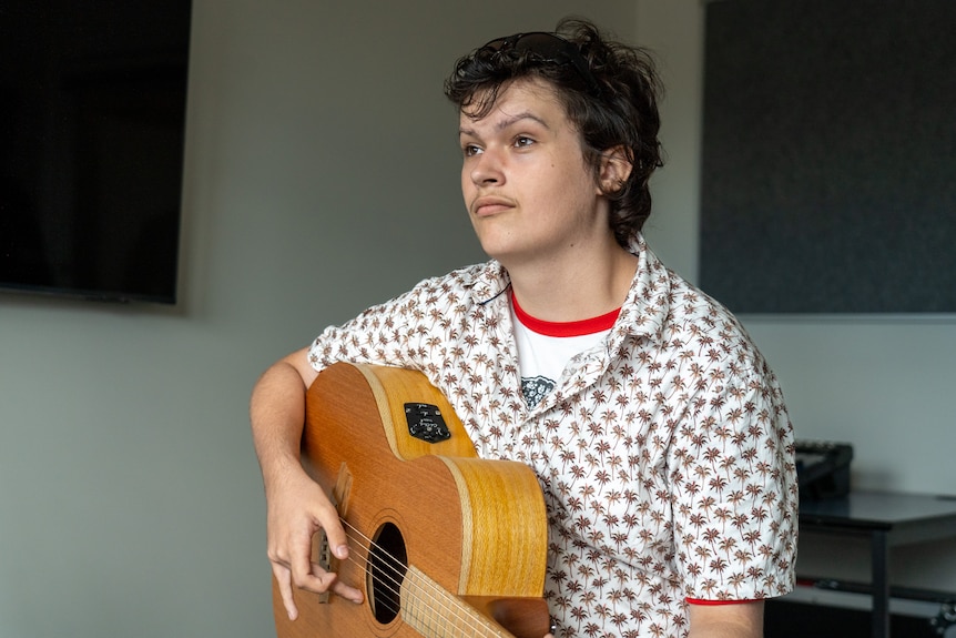 a young man holding an acoustic guitar looks pensive