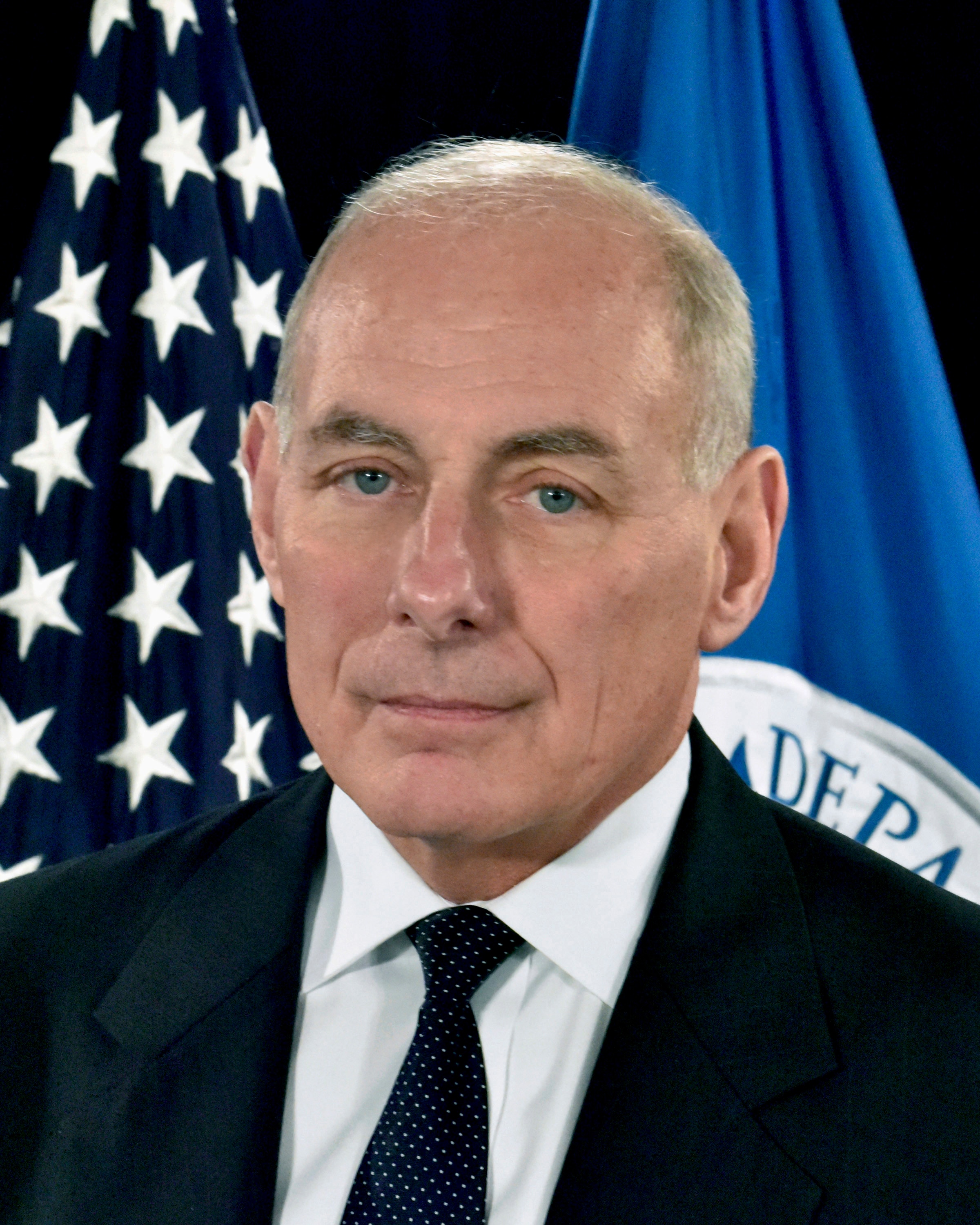 A portrait of John Kelly, who wears a black suit and dark tie.