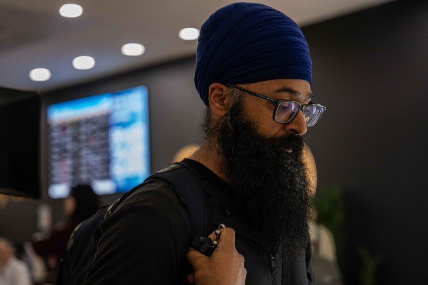 A man with a long beard, turban, and glasses looks down as he walks indoors. He is wearing a backpack.