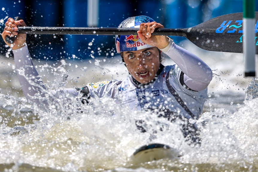 Kayaker Jessica Fox competing, holding her paddle above her head, going through a gate, water splashing over her