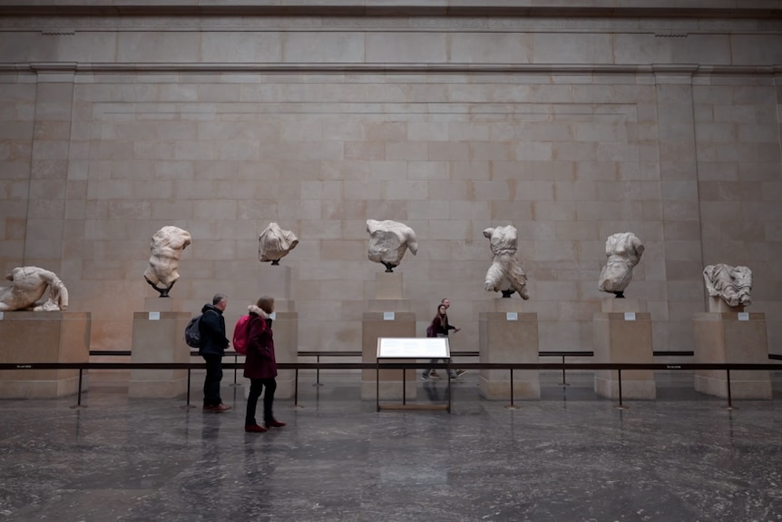 A wide shot of marble statues, many damaged, on display in a row in a big hall in the British Museum