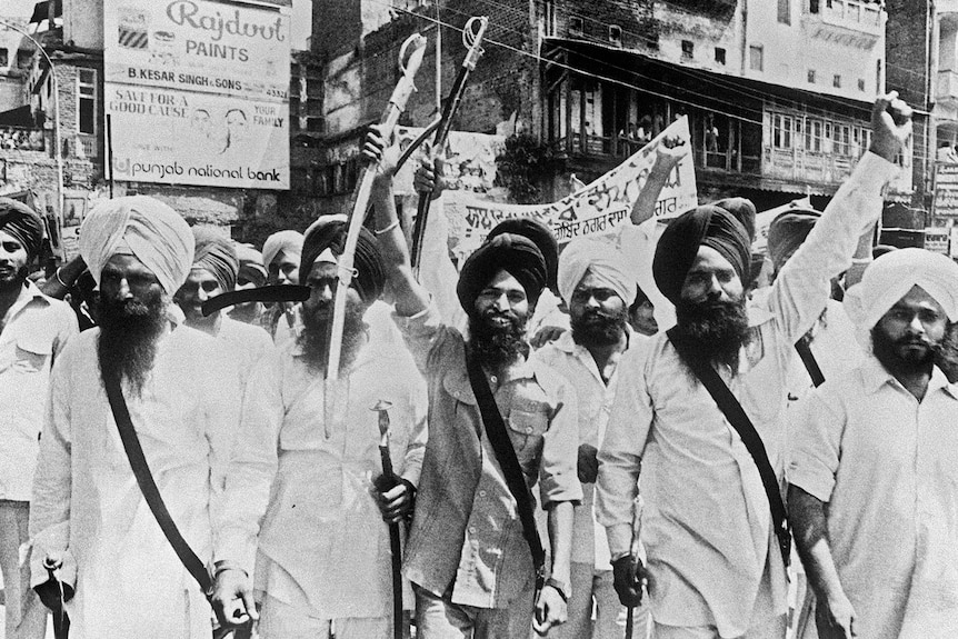 A black and white photo of SIkh men wearing turbans marching on a street several hold sheathed swords above their heads.