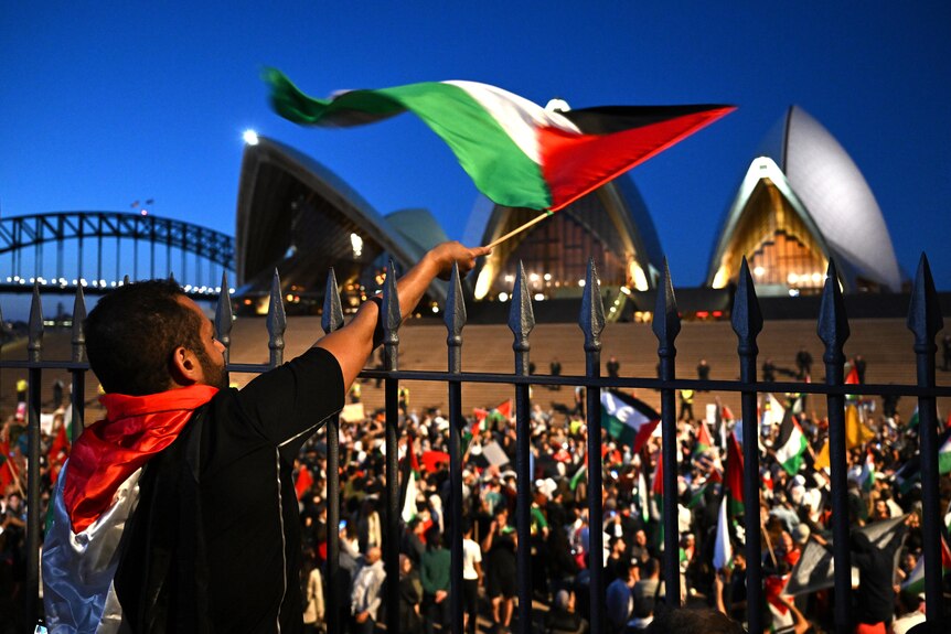 A man waves the Palestinian flag in front of a crowd gathered on the steps of the Sydney Opera House.