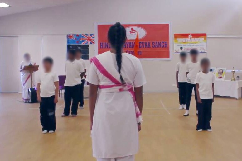 Boys stand in formation in a hall. In front of them stands a woman, her back to the camera.