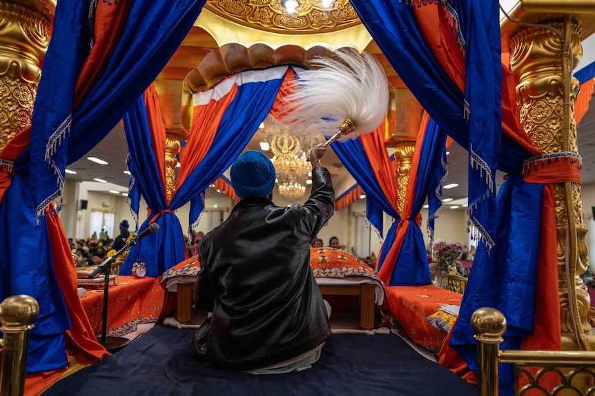 A man sitting on gold platform adorned with blue and orange fabric waves a tassled object above his head.