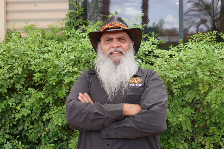 A man wearing a hat with a long white beard smiles in front of a green hedge.