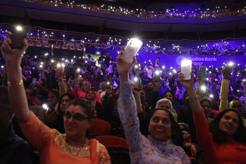 Hundreds of people hold their phones up with their torches on in a darkened arena. Some in the front can be seen smiling