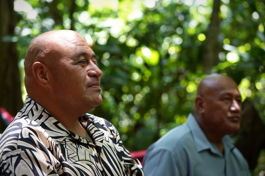 Two men look off camera, with trees in the background. 