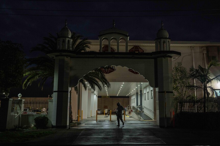 A Sikh place of worship is partially lit at night. A person walks past it silhouetted against the light.