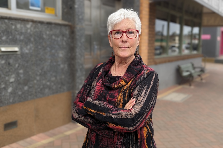 A middle aged woman wearing a sombre colourful top stands in front of a granite block building. 