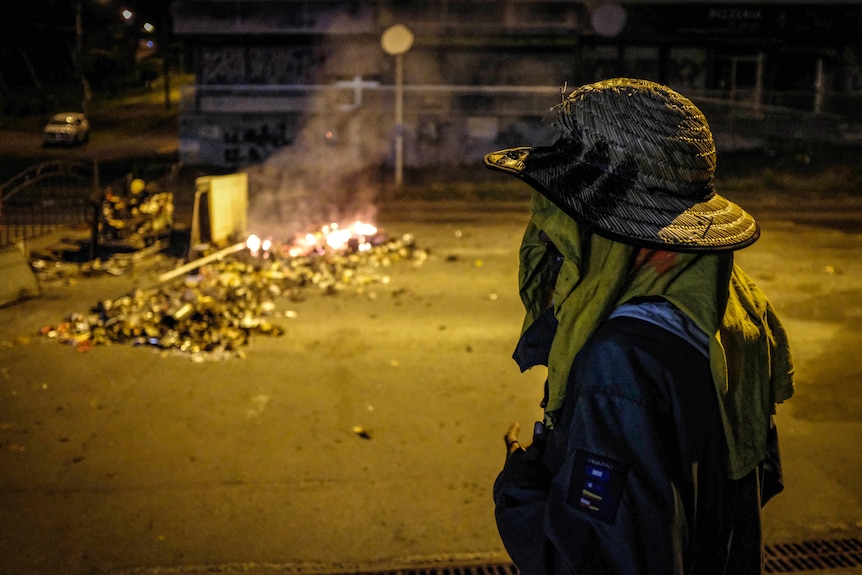 A man concealing his face stands in front of a roadblock and a small fire at night.