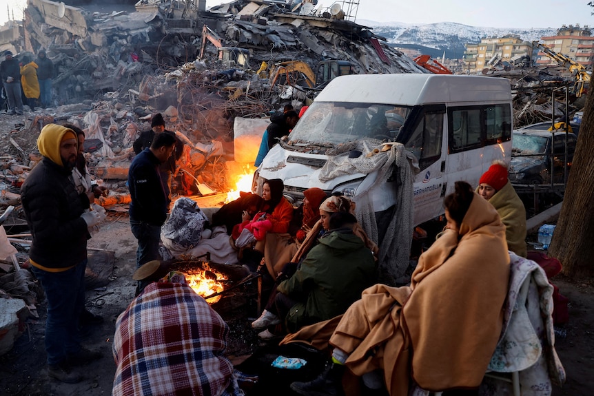 People sit around a fire next to rubble and damages near the site of a collapsed building in the aftermath of an earthquake