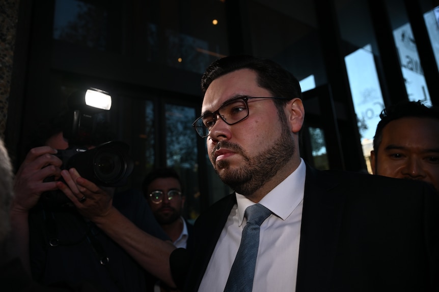 A man walks out of glass doors flanked by a lawyer and journalists.