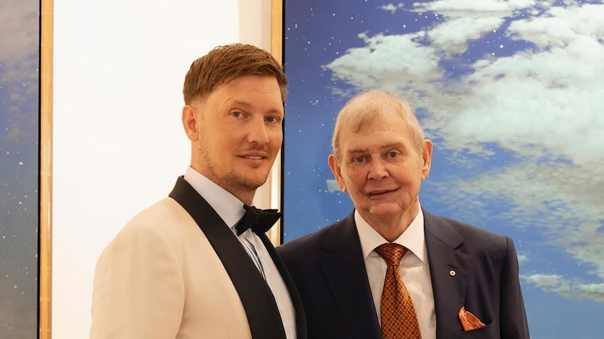 Singer John Farnham standing next to his son Rob at his wedding. John is wearing a black suit while rob is wearing a cream suit