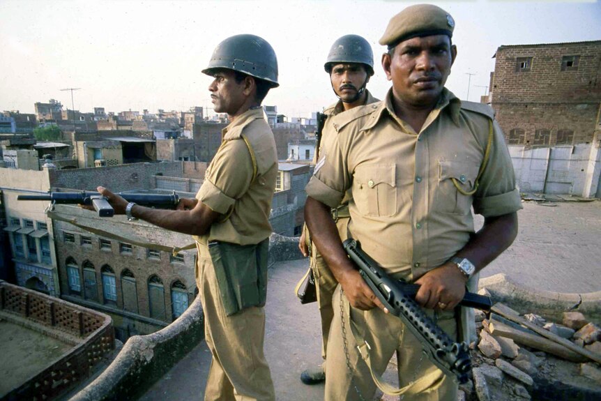 Three men in Indian army uniforms, two holding machine guns, stand on a rooftop.