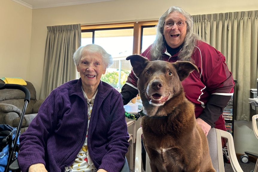 two women with greyish/white hair and a dog sitting up on a chair smile at the camera