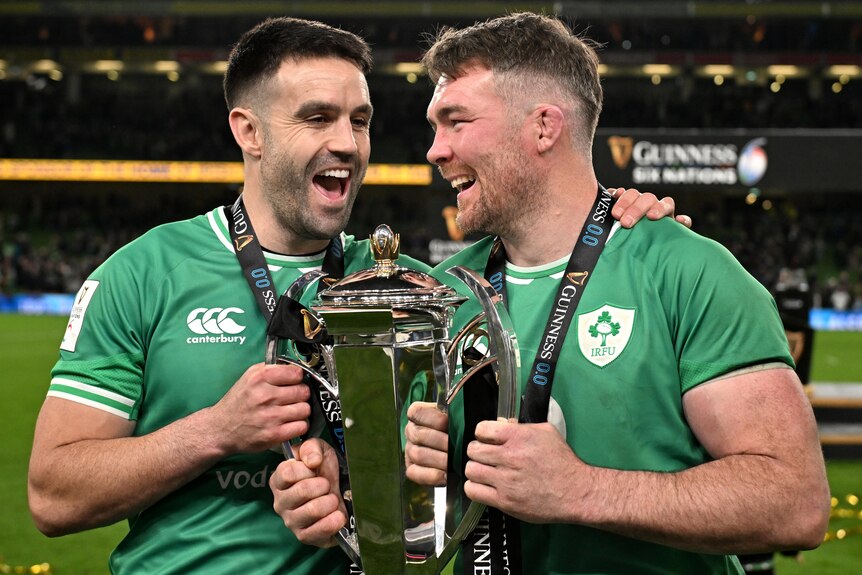 Two Ireland players smile as they hold the Six Nations trophy.