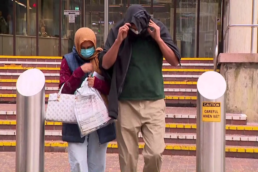 A man and a woman who are covering their faces with masks and a jumper walk from a court building. 
