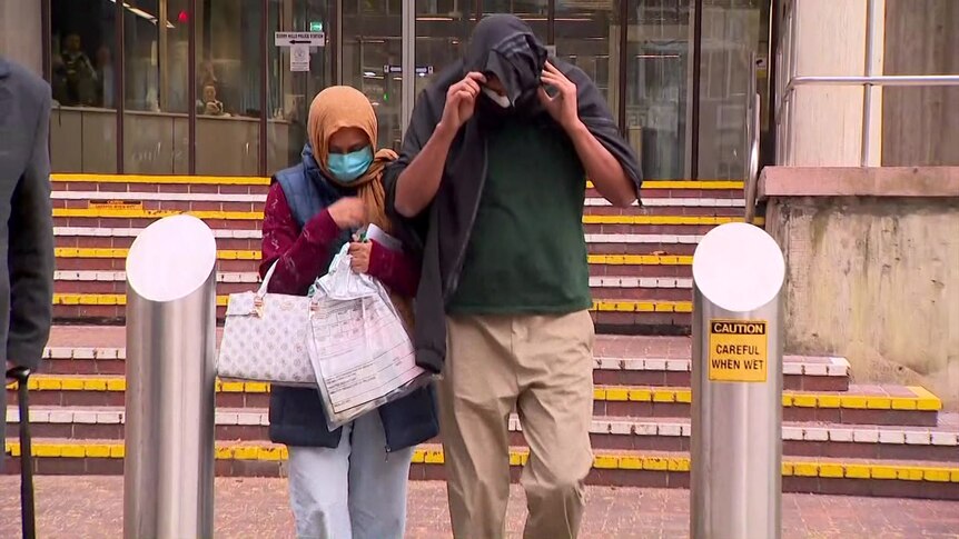 A man and a woman who are covering their faces with masks and a jumper walk from a court building. 