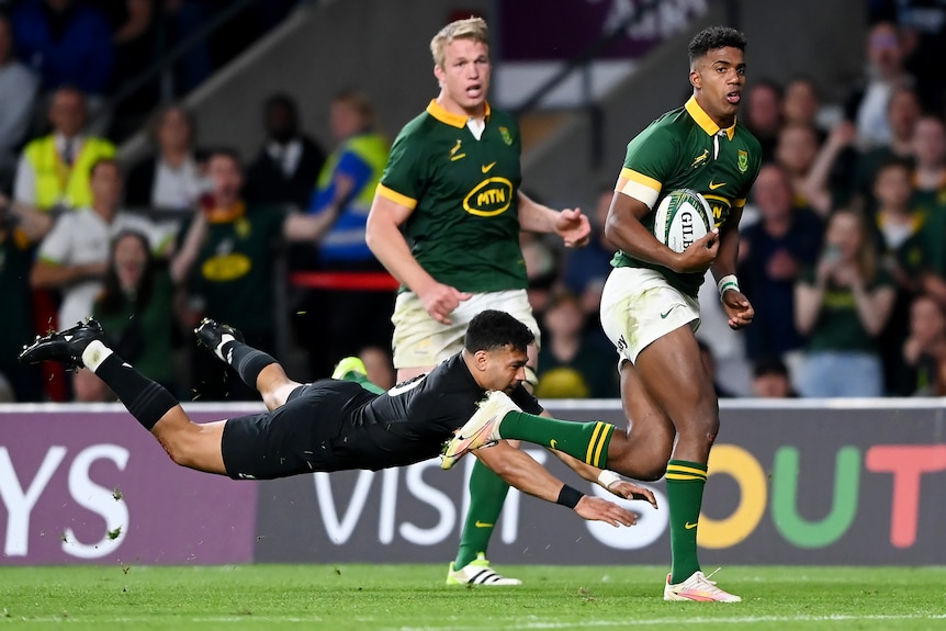 A Springboks player runs with the ball in his right hand as an All Blacks opponent dives to in an attempt to tackle.