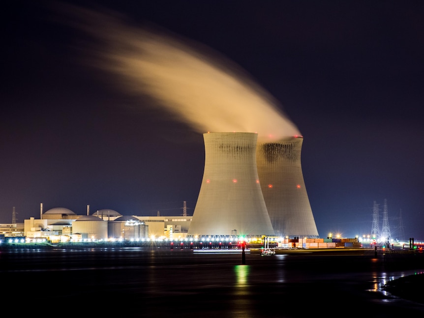 A long exposure shows a nuclear plant illuminated at night, with a white cloud of steam emerging from the top of two reactors. 