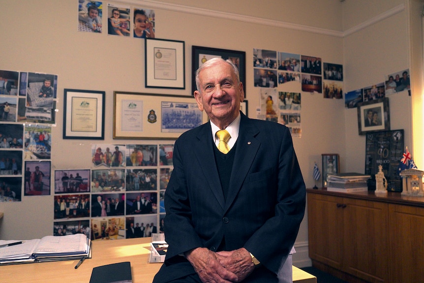 An older man sits on the edge of a desk in an office, smiling and wearing a suit and yellow tie. Photos adorn the wall behind