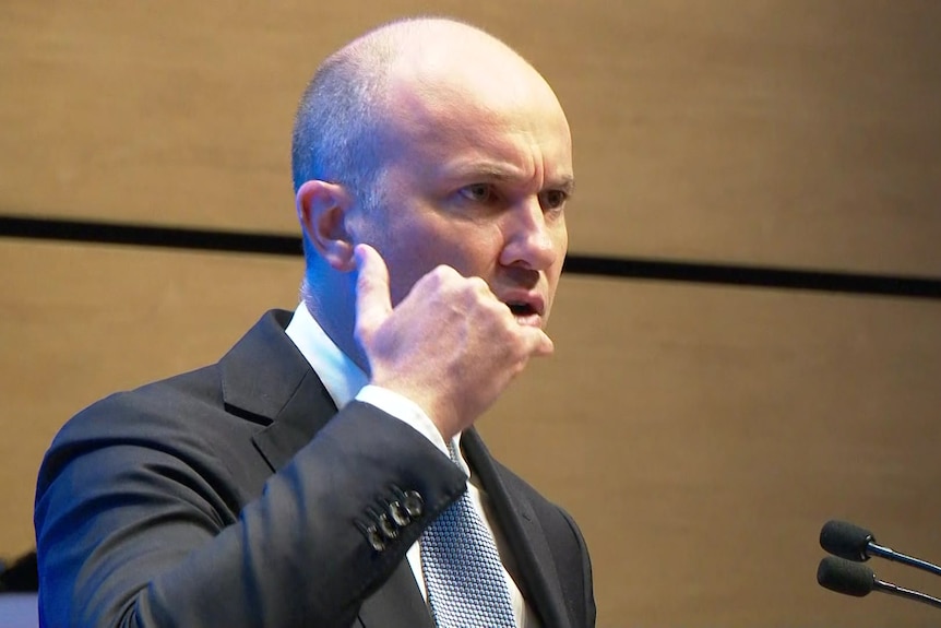 a man standing at a podium with his hand up against his ear pretending to speak into a phone