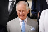 King Charles III smiles while wearing a grey suit with a blue tie with the Greek flag and a matching handkerchief