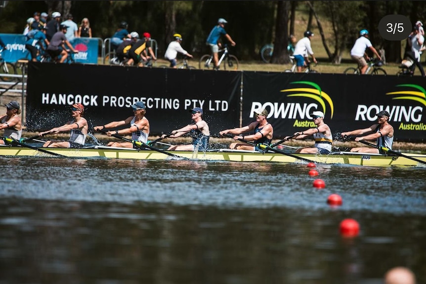 A wide shot of a group of Australian rowers on a boat on the water with cyclists visible in the background on land.