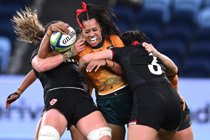 A Wallaroos player holds the ball while being tackled by two Canadian players.