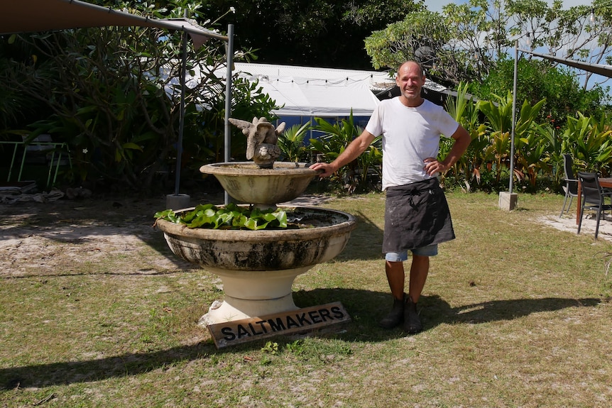 A man stands in a garden with his hand resting on a stone fountain with the sign "Saltmakers".