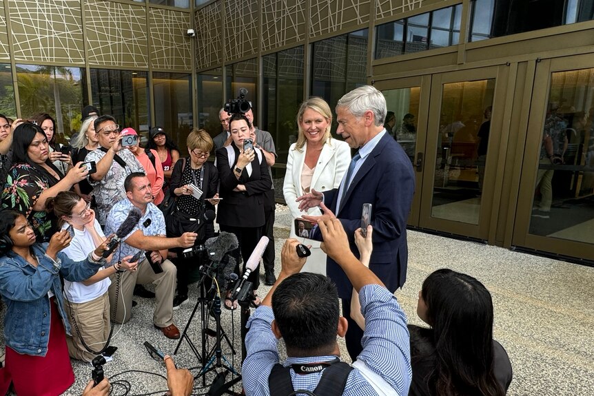 A woman wearing white smiling while standing next a man in front of a group of people with cameras and microphones