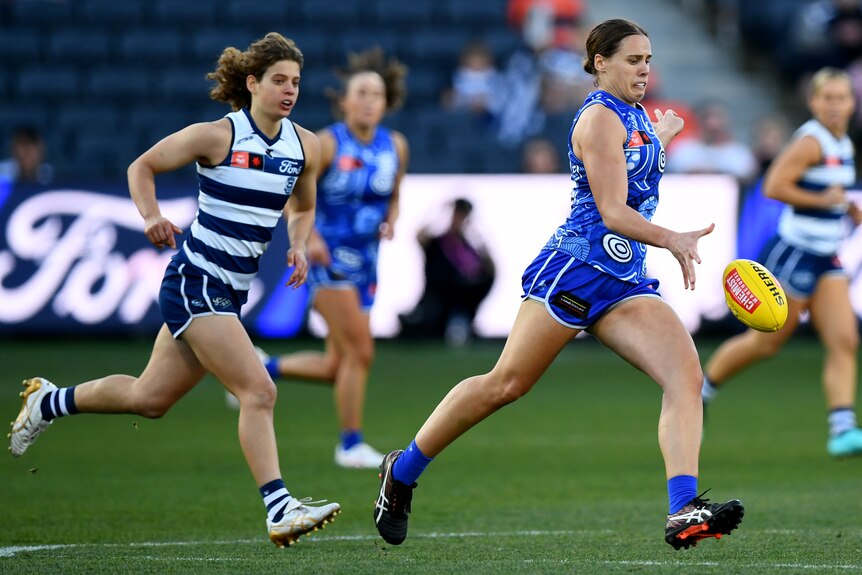 A Kangaroos AFLW player runs clear in the middle of the ground as she drops the ball to kick it as a defender trails. 