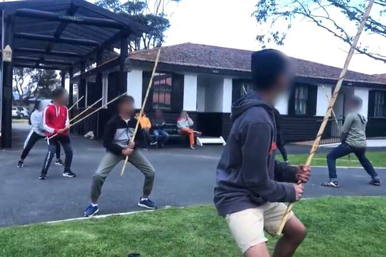 A number of teenage boys stand outdoors in a ready stance, holding large bamboo sticks.
