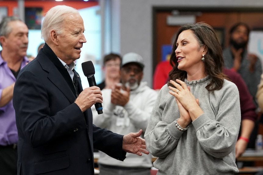 Joe Biden holds a microphone and speaks. Gretchen Whitmer stands next to him and claps her hands.