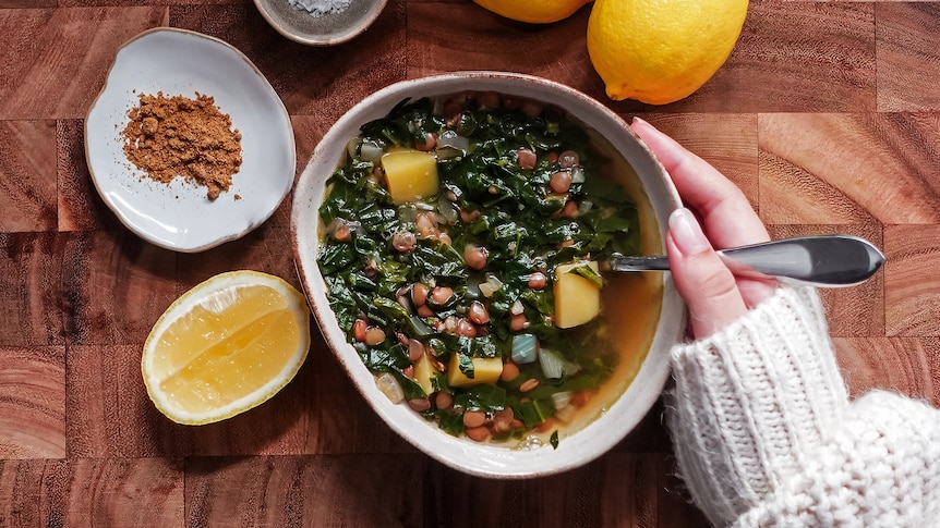 A hand hugging a bowl of lentil soup with potato and spinach. On the table are lemons, cumin and sea salt.