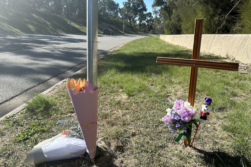 A grass strip next to a road with multiple bouquets of flowers and a wooden cross.