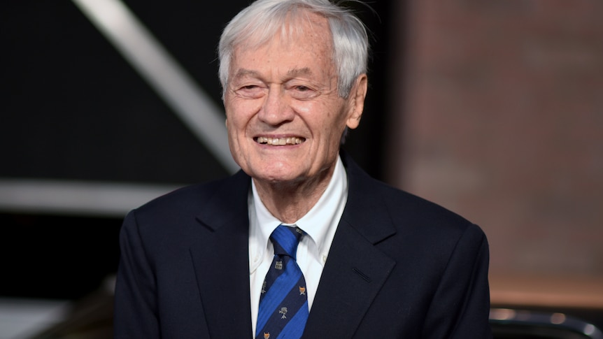 A grey haired man in a suit with a blue neck tie smiling. 