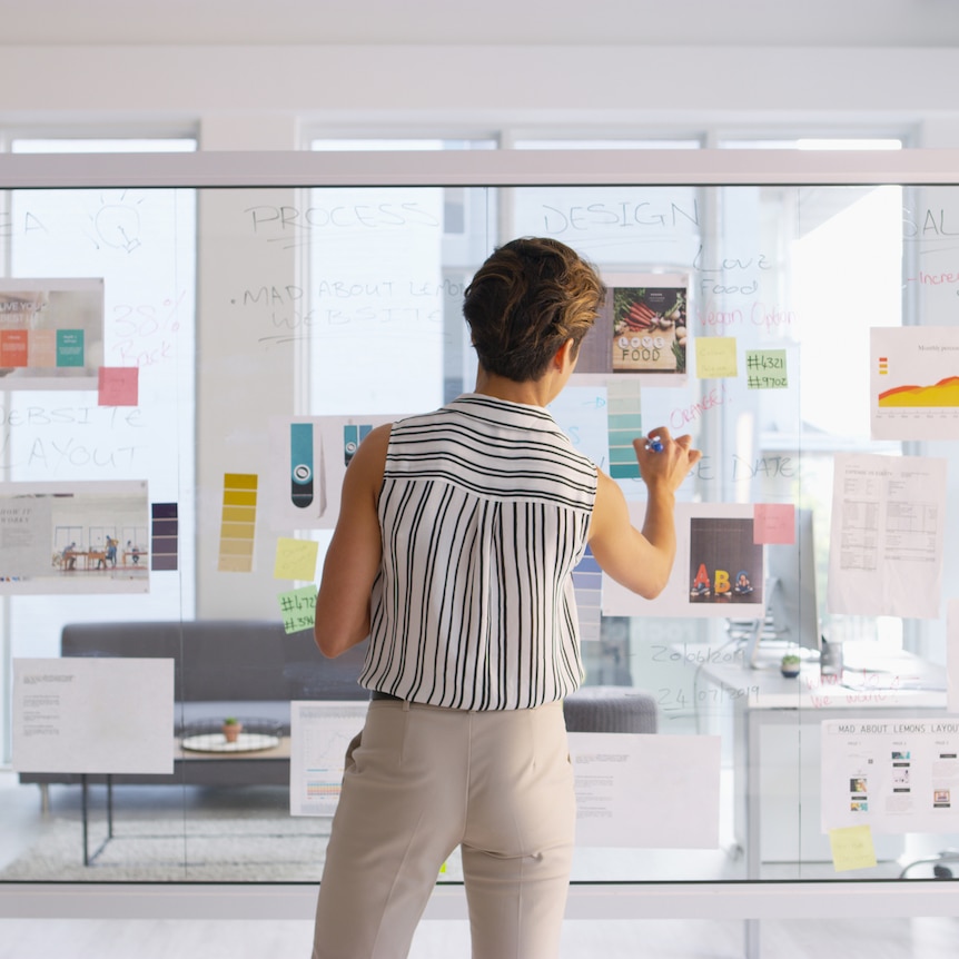 woman in modern office brainstorms on glass wall with marker and post it notes