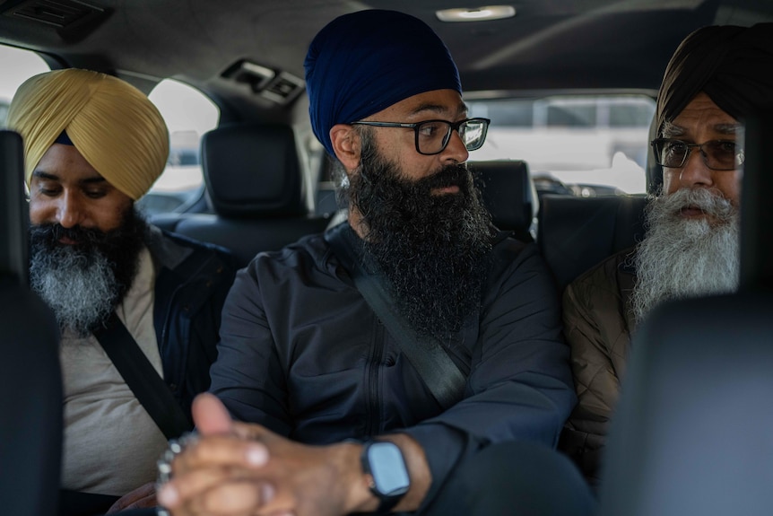 Three men wearing turbans and beards sit in the back of a car. The man in the middle is looking to the side.