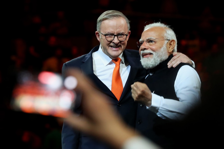 Anthony Albanese smiles as he puts his arm around Narendra Modi. Modi is making a fist. The background behind them is dark.
