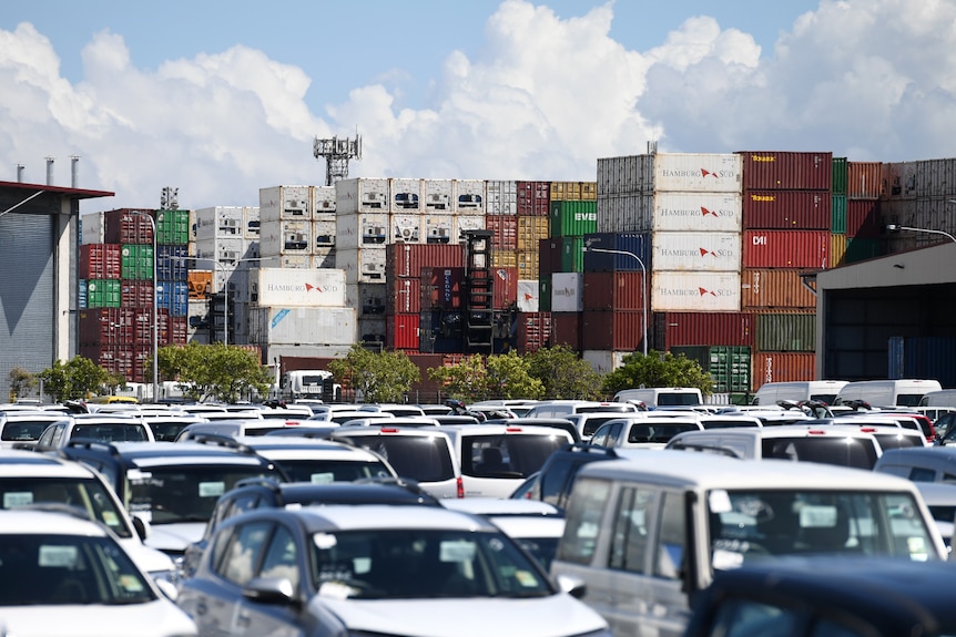 Rows of cars in a car park, with stacked shipping containers behind them.