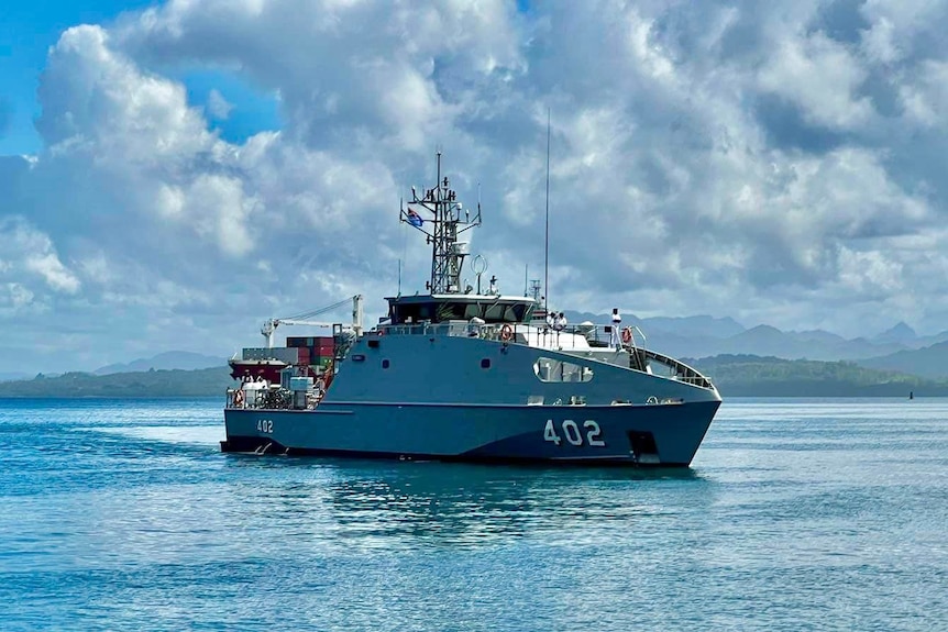 A grey navy patrol boat sails into a harbour on a cloudy day, with mountains in the background.