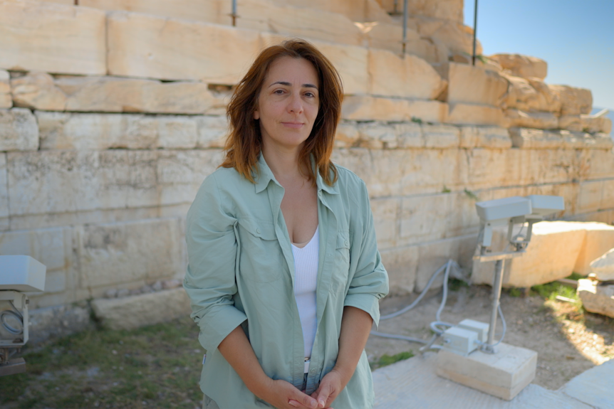A woman with brown/red hair wearing a light green long-sleeved shirt stands in front of a large stone ruin in the shade