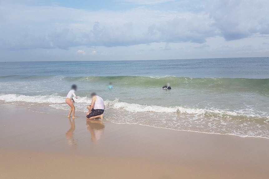 A man and a young girl crouch down on the sand at the edge of the ocean, while two people can be seen in the water