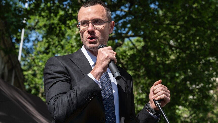 A man speaks into a microphone at a rally in a UK park.